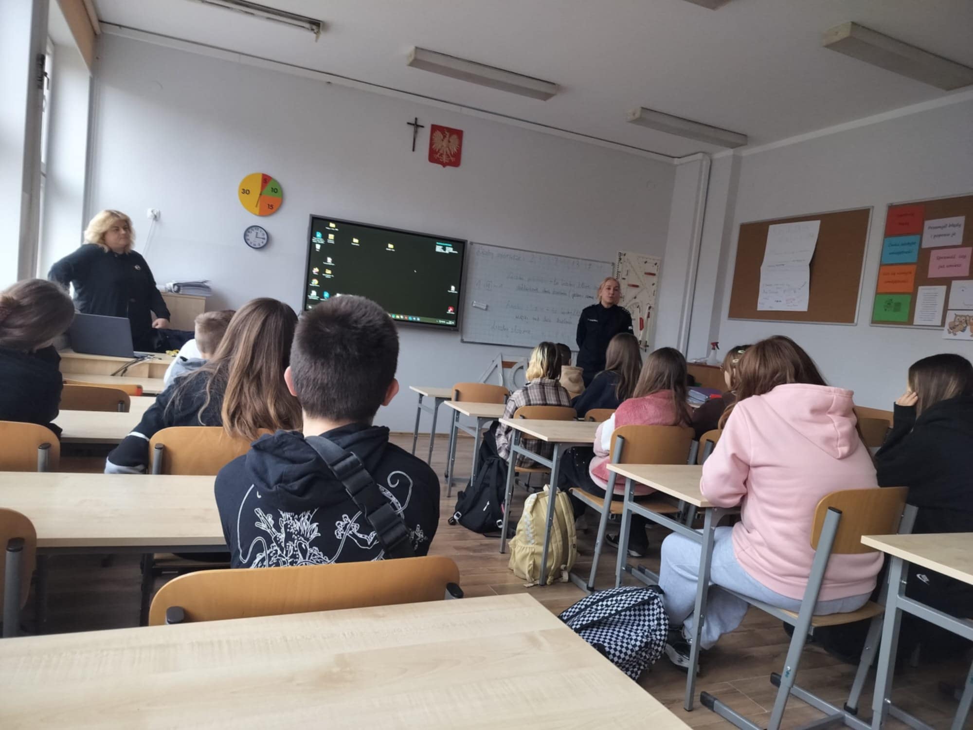 Uczniowie siedzą w ławkach szkolnych, mówi do nich pani policjantka.